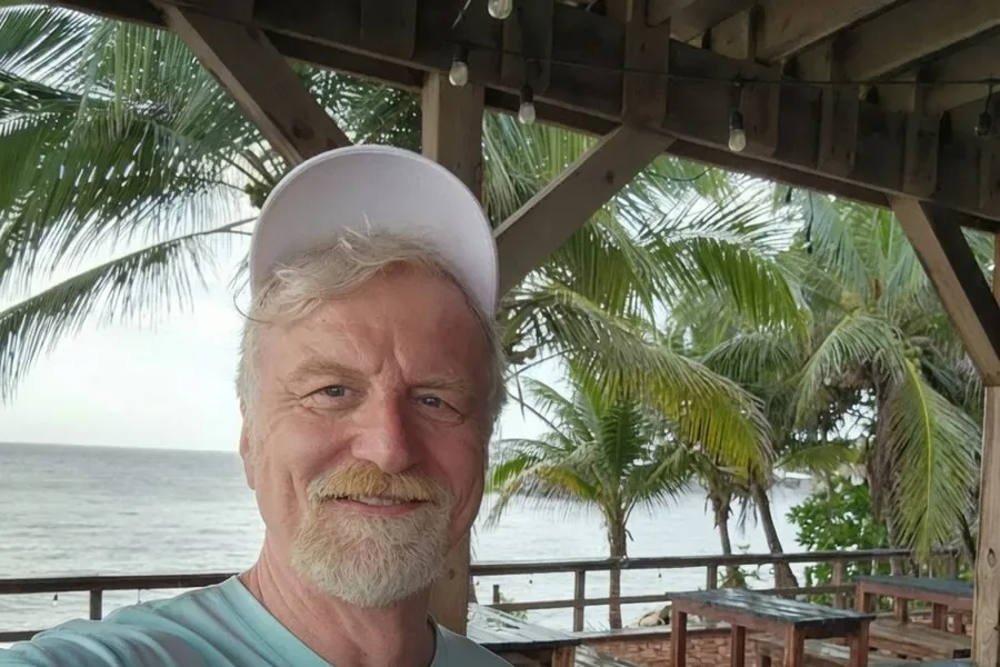 Andrew Easy Anderson at a beachside pavilion with palm trees and the ocean behind him.